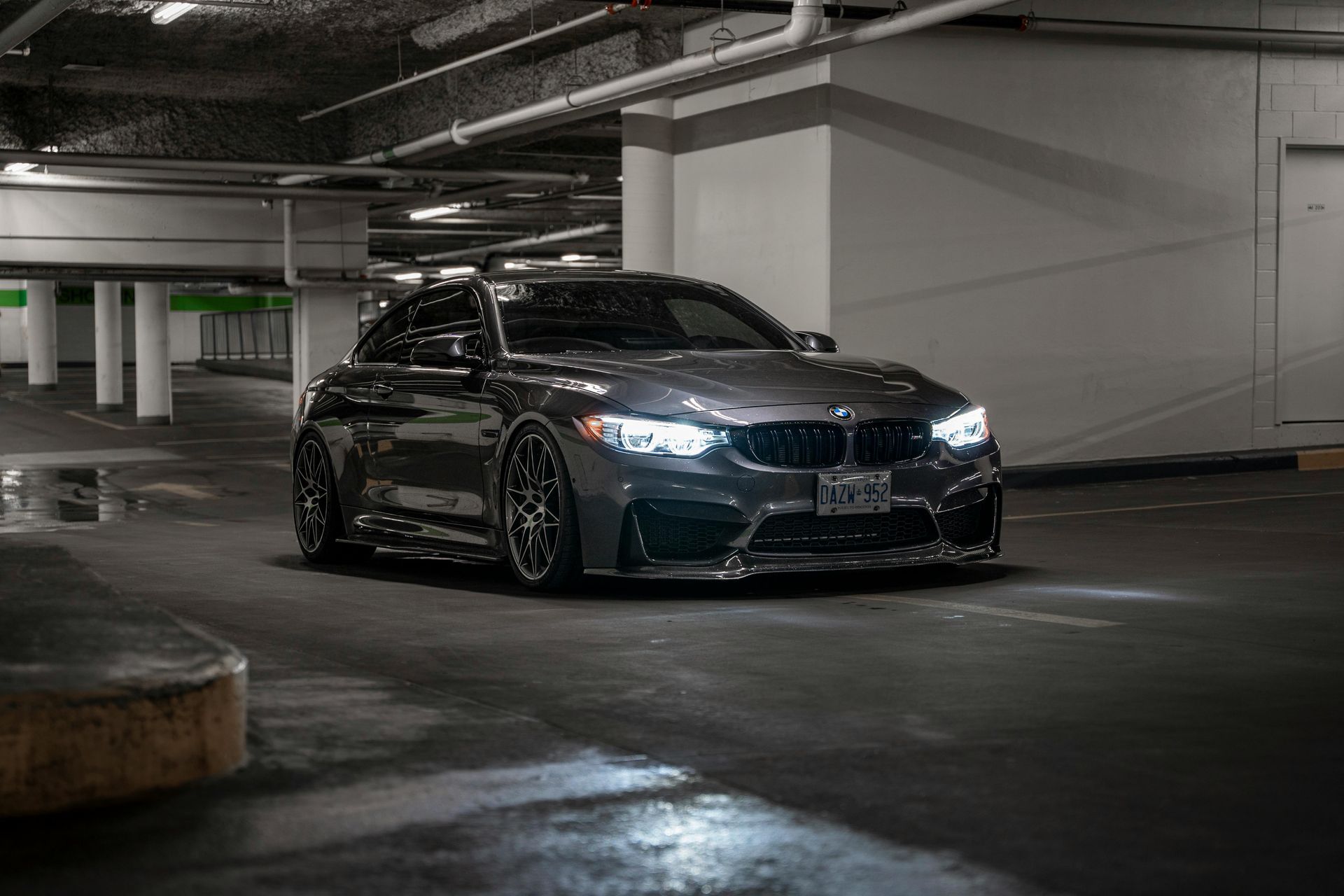 Dark gray BMW M4 sports car in a dimly lit parking garage, headlights on.