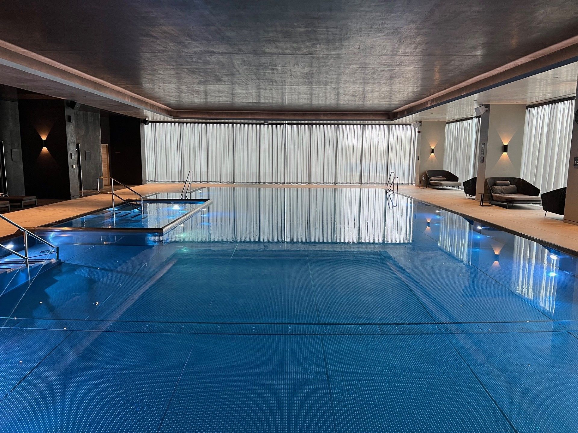 Indoor pool with blue water and silver ceiling.  White curtains in the background, seating areas on either side.