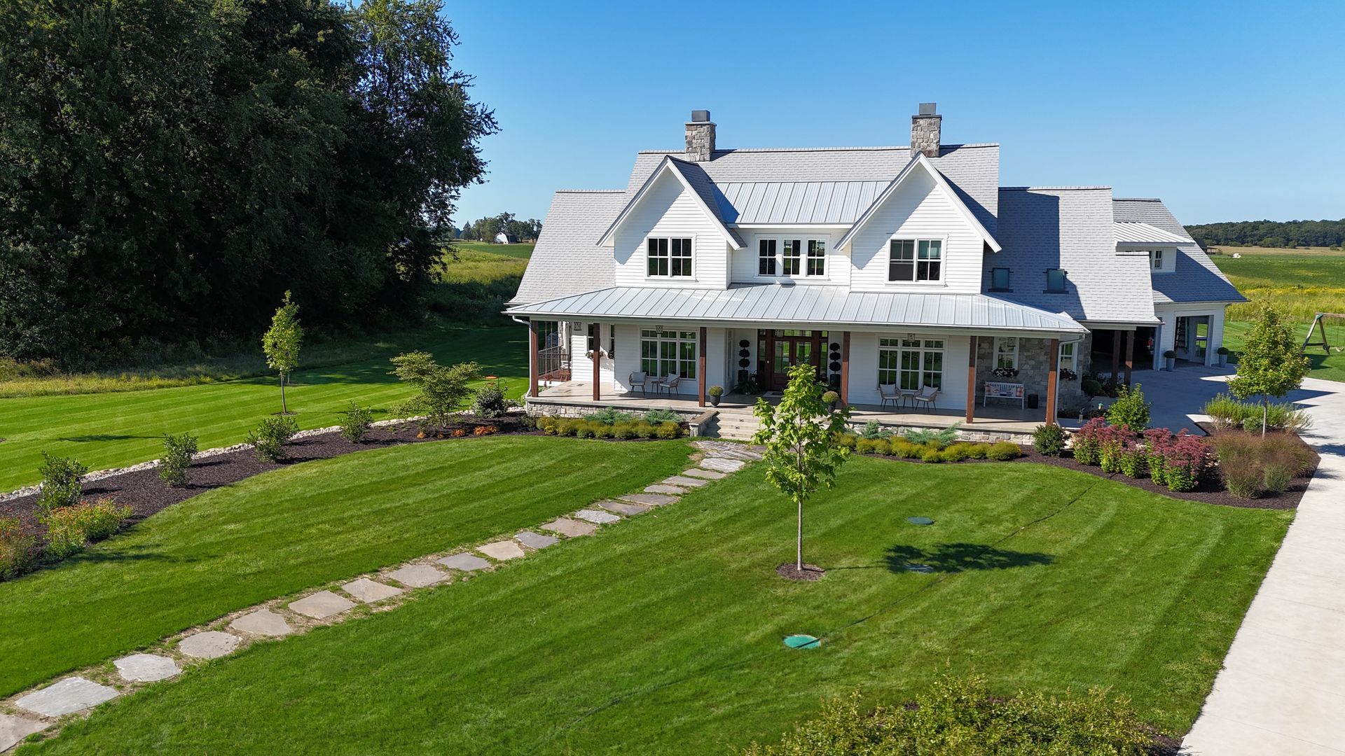 White farmhouse with wrap-around porch, gray roof, and well-manicured lawn on a sunny day.