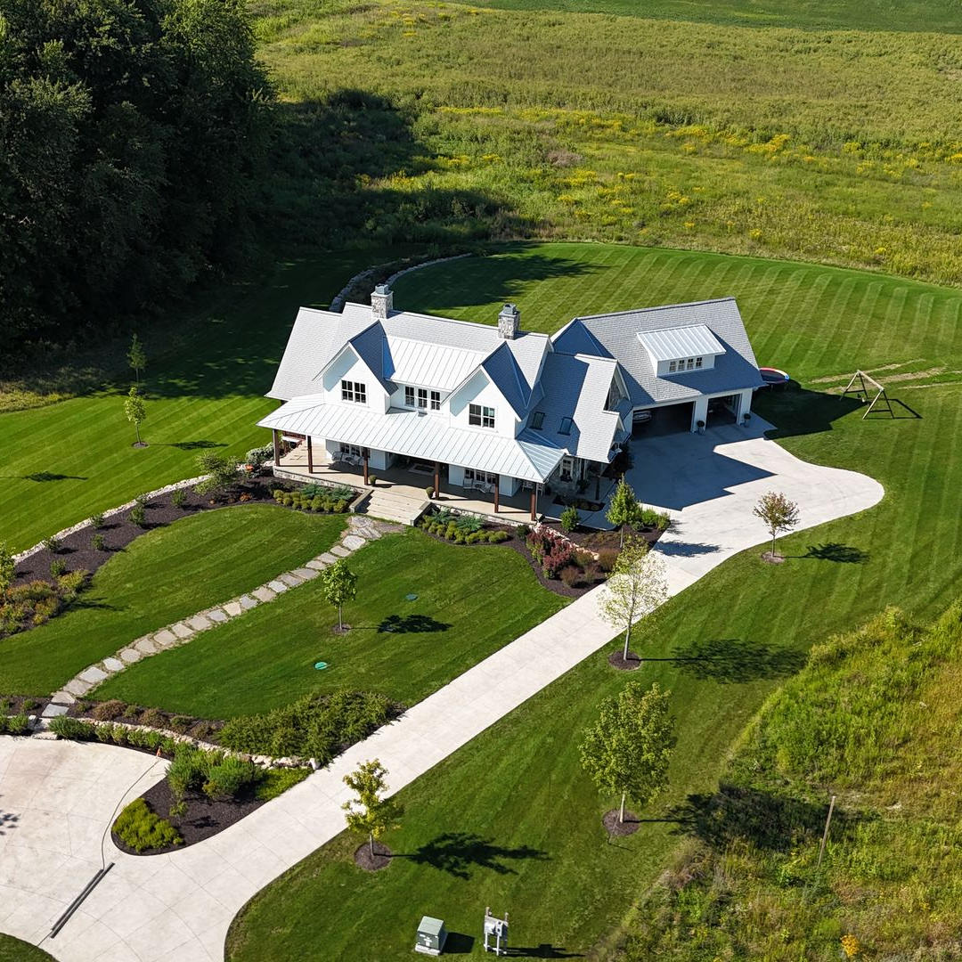 White farmhouse with a gray roof on a large, green lawn under a blue sky.