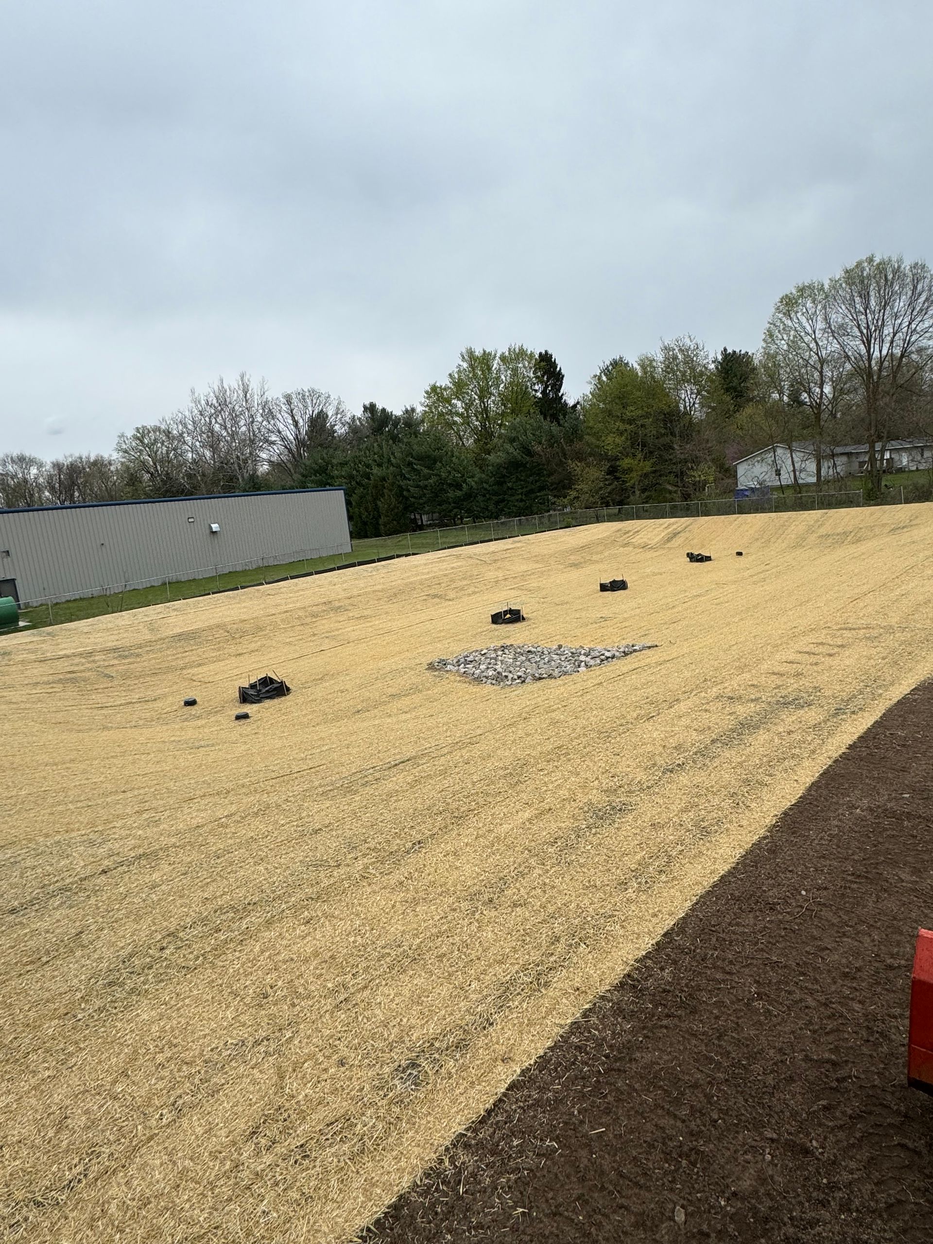 A sloped landscape covered in tan mulch and scattered rocks, under a cloudy sky.