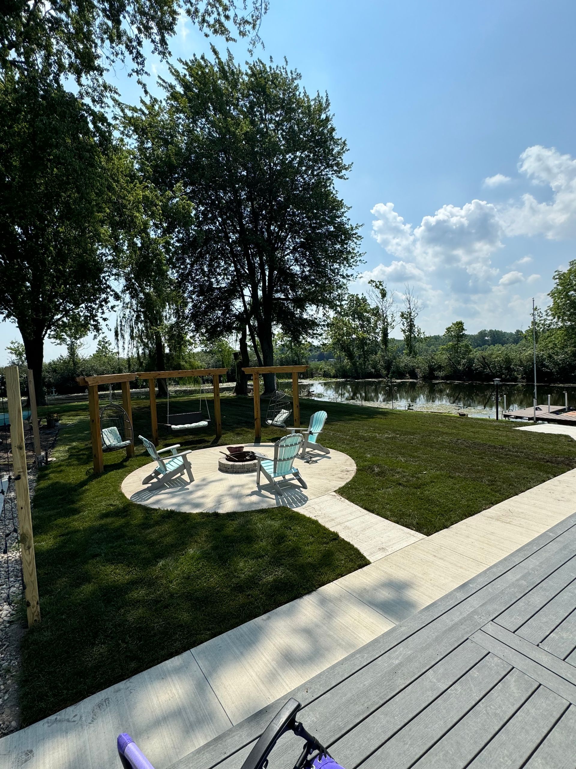 Lakeside fire pit area with light blue chairs, concrete patio, grass, trees, and wooden swing set.