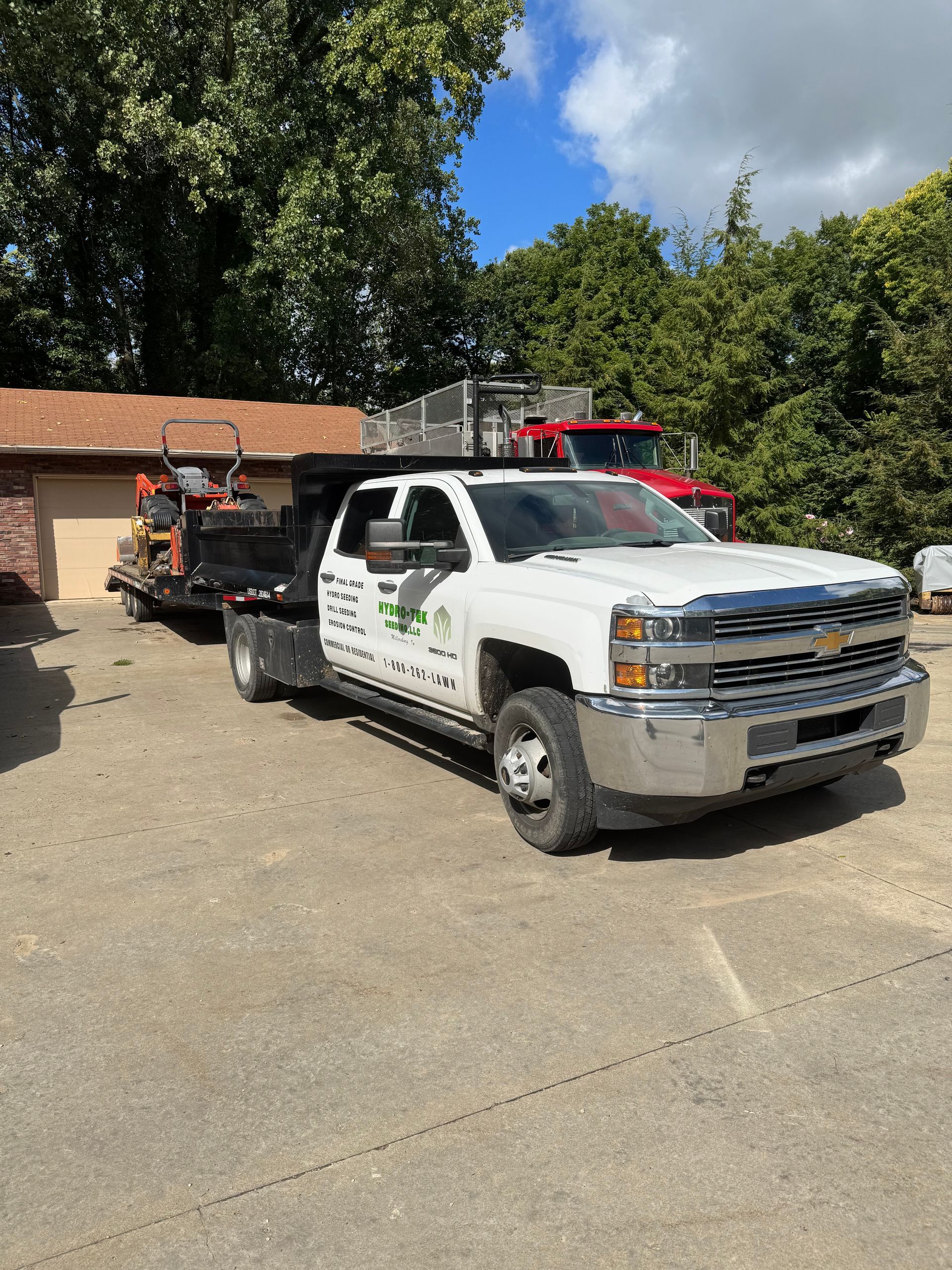 White landscaping truck pulling a trailer with a mower, parked outside a building on a sunny day.