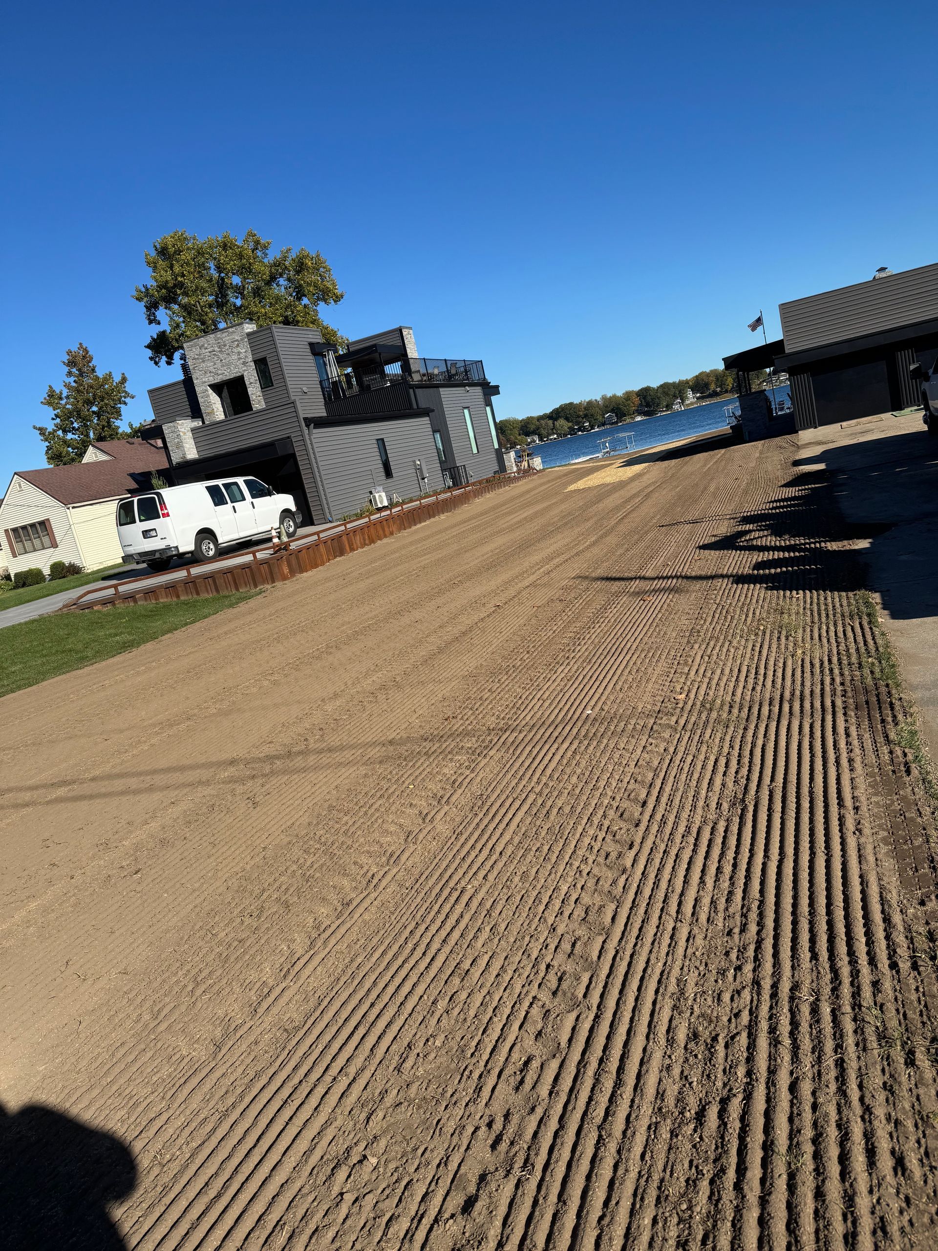 Gravel driveway leading to buildings and lake under a blue sky. A white van is parked.