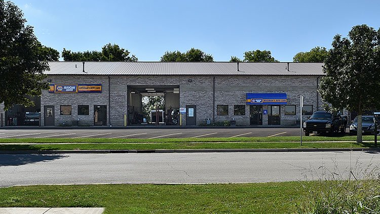 Single-story strip mall with storefronts, parked cars, and trees along a road.