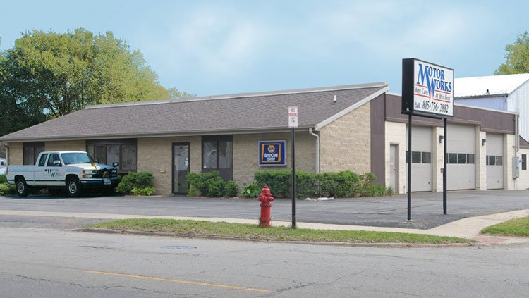 Exterior view of a motor works shop with a white truck parked out front; blue and white sign, beige brick building.