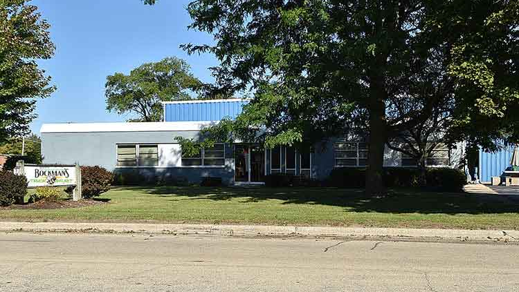 Low white building with blue roof behind trees and lawn beside a street, with a sign at left