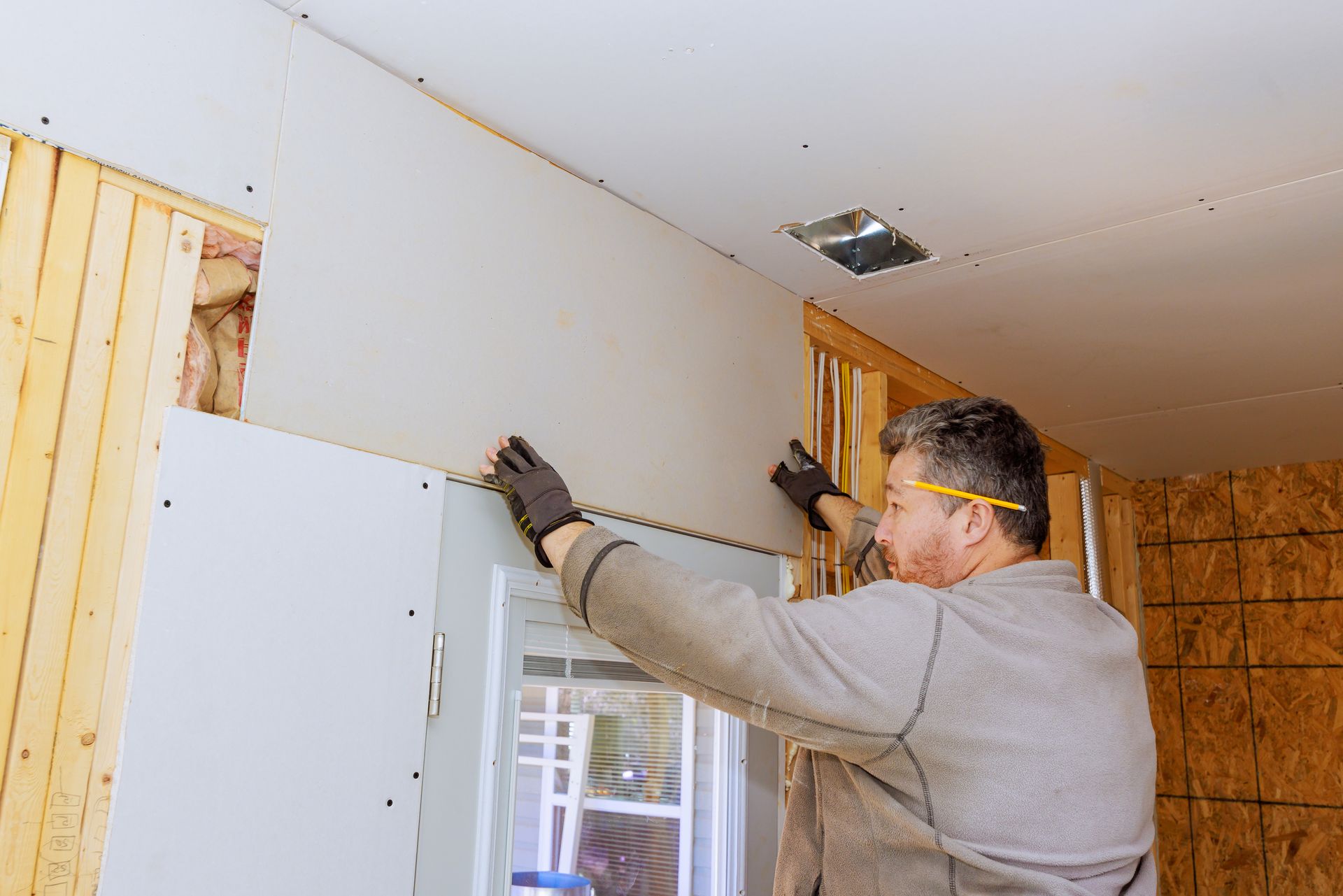 A man is installing drywall on a wall next to a window.