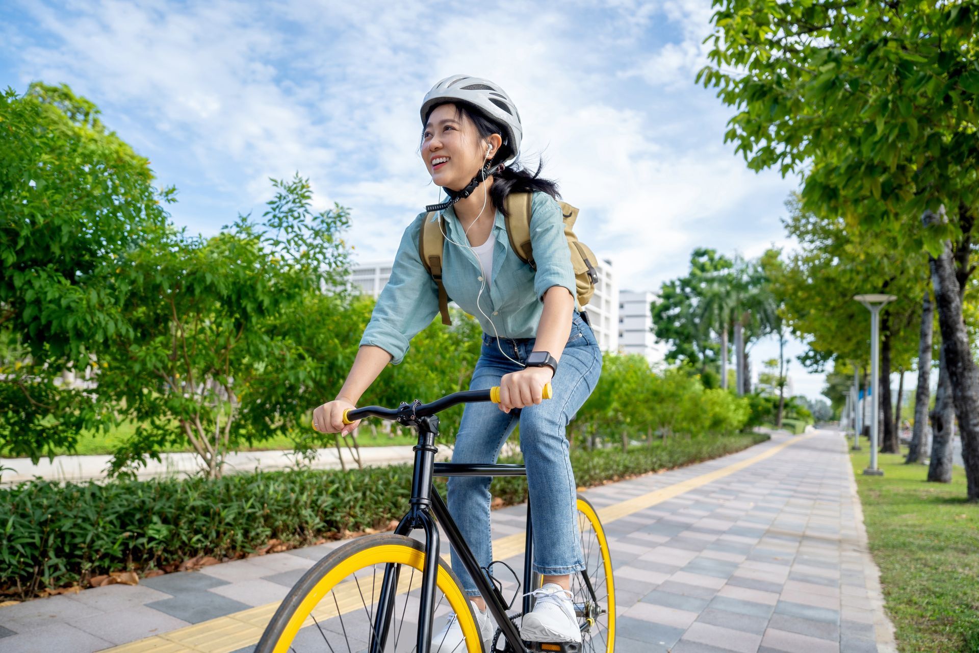 A person wearing a helmet and backpack rides a bicycle with yellow rims along a paved path lined with trees.