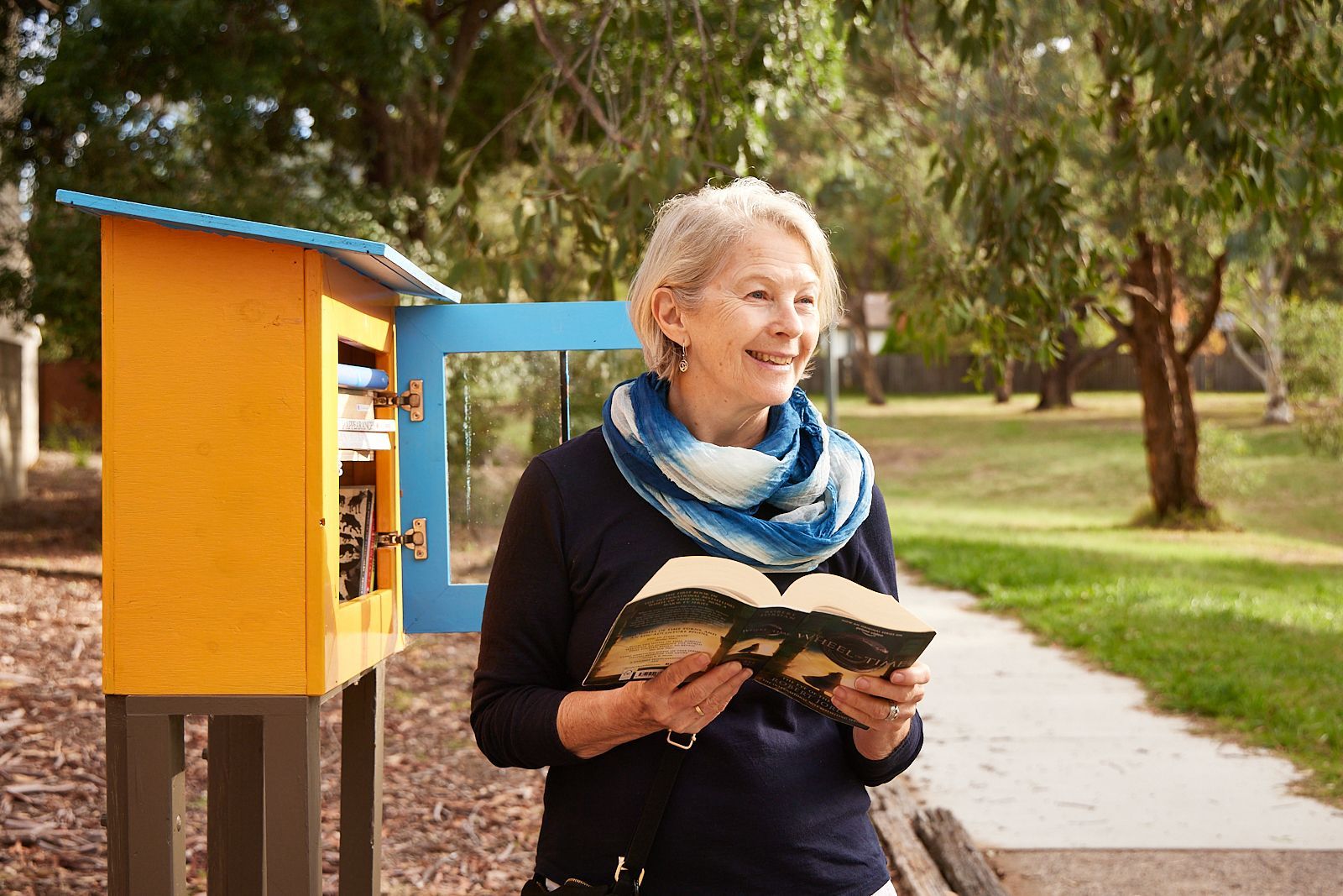 A person smiling while holding an open book near a small, bright yellow roadside library in a park.