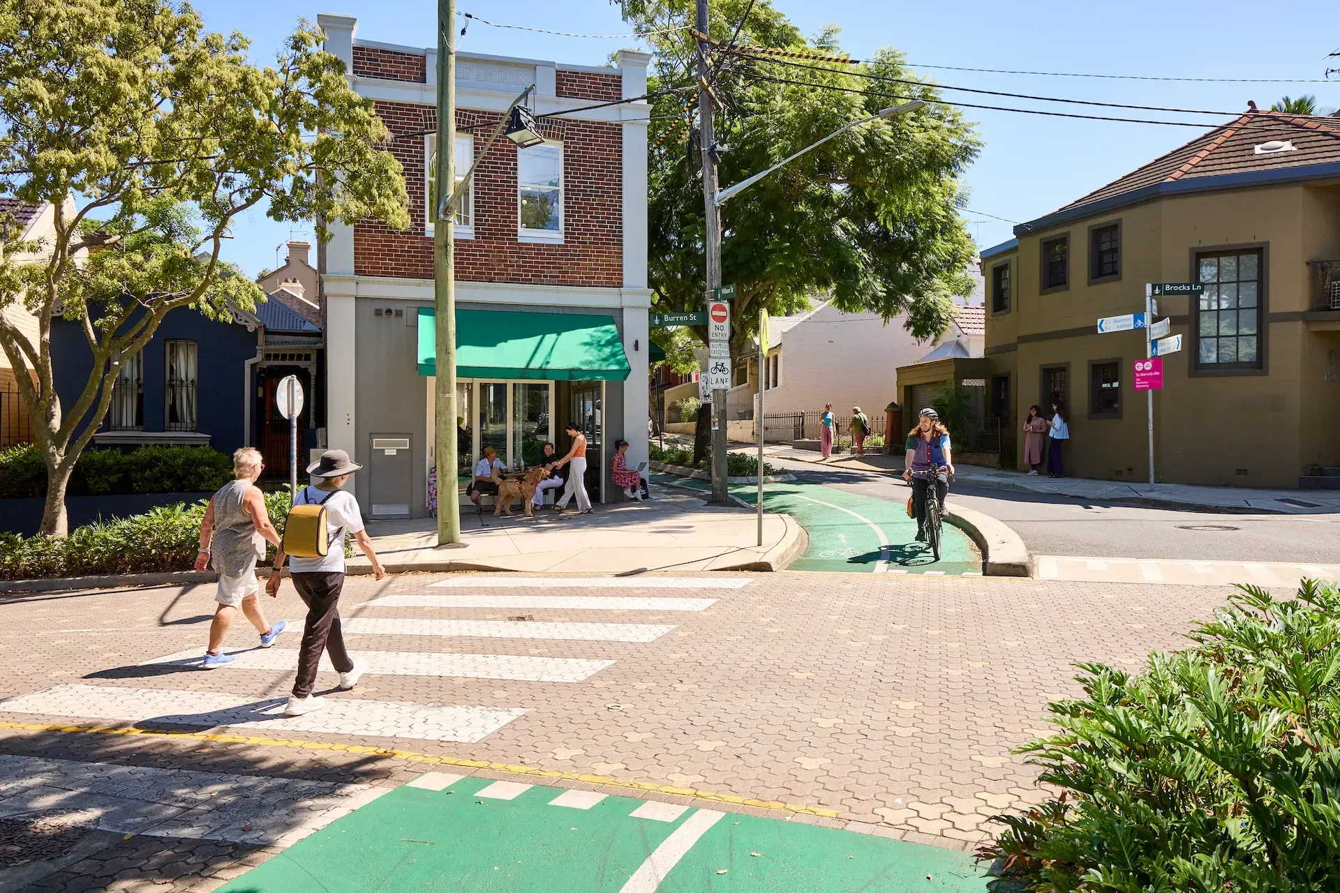 A sunny street intersection featuring a brick building with a green awning, a crosswalk, and a green painted bike path.