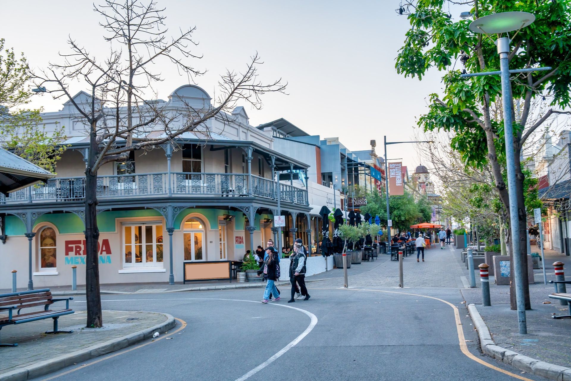Pedestrians cross a street lined with shops and outdoor seating, featuring a prominent corner building with a balcony.