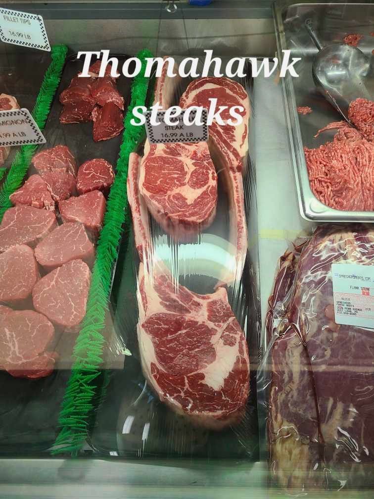 Tomahawk steaks displayed in a grocery meat case with other cuts on green trays.