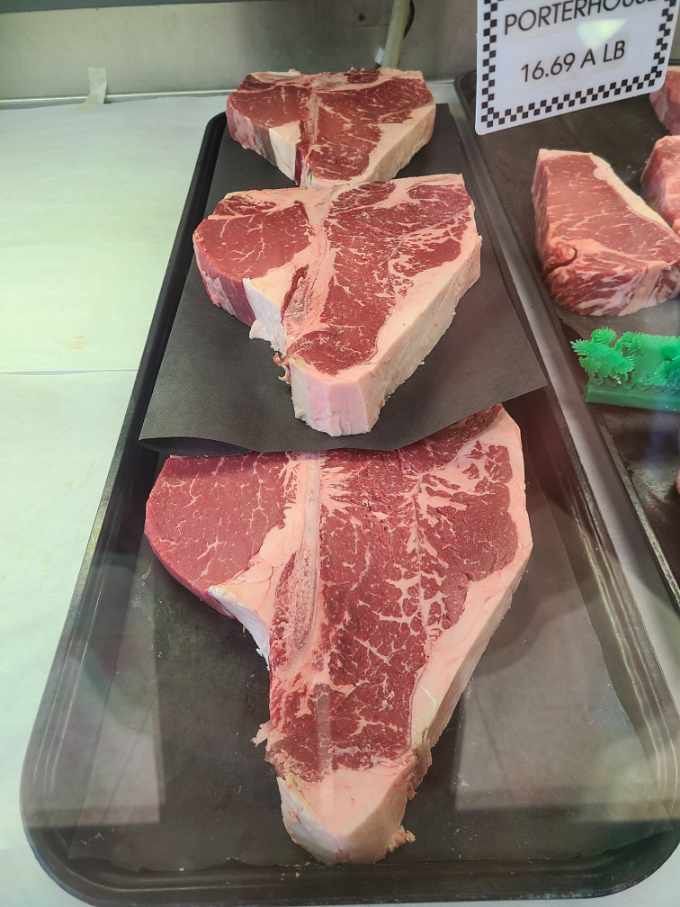 Raw porterhouse steaks displayed on a black tray at a butcher counter, with a price sign nearby