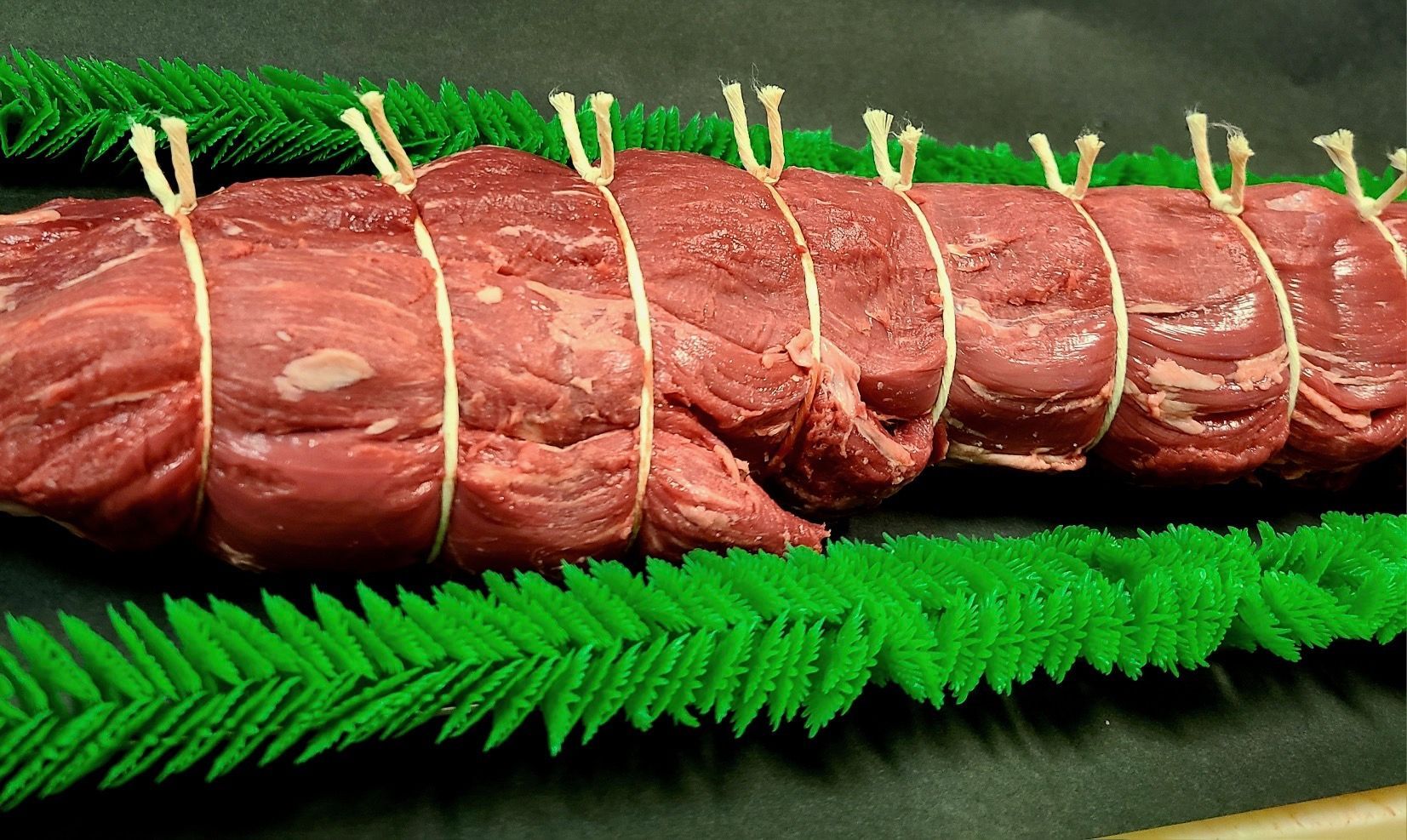 Raw beef roast tied with twine, resting on bright green fern leaves on a dark platter
