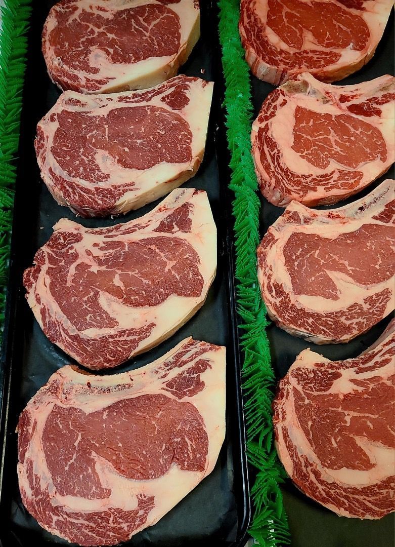 Six raw marbled steak cuts arranged in two vertical rows on black trays with green garnish.