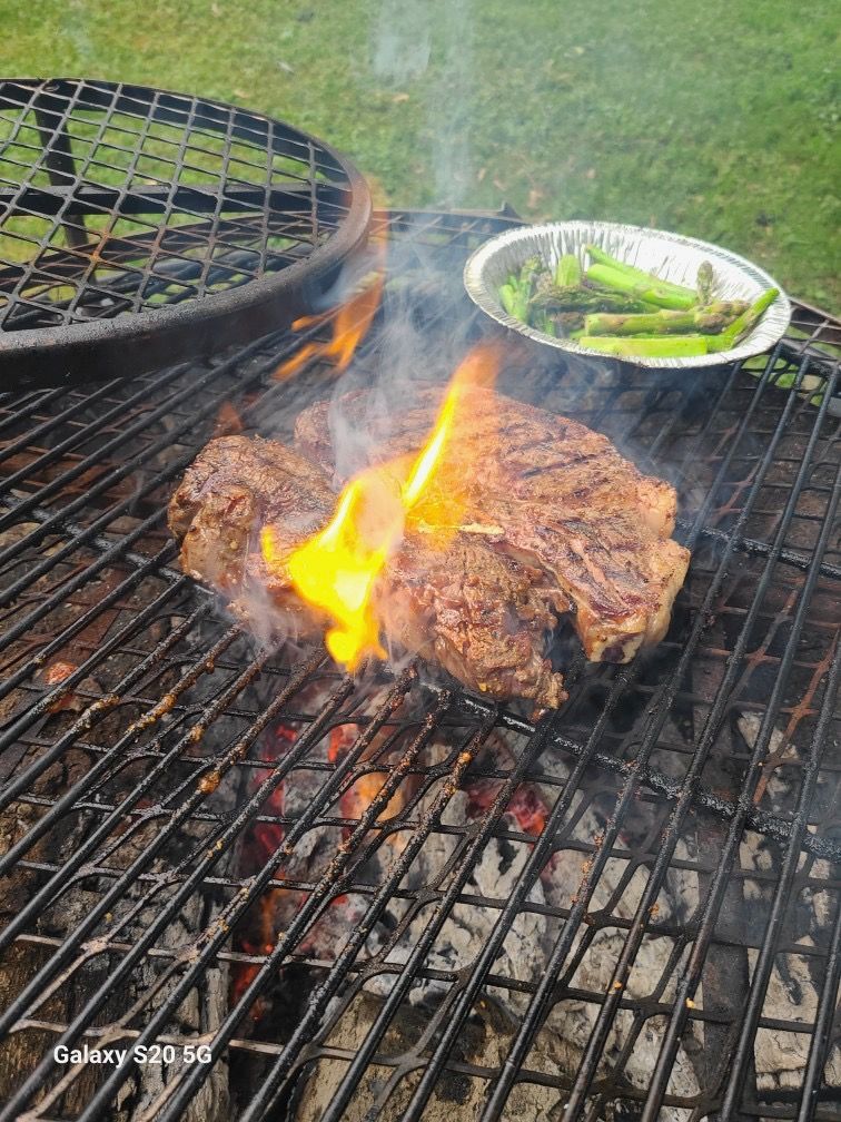 Steak grilling over flames on a barbecue, with a tray of green vegetables nearby.
