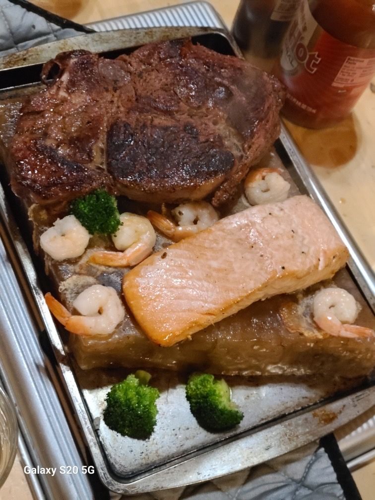 Tray of grilled salmon, steak, shrimp, and broccoli with sauce on a table