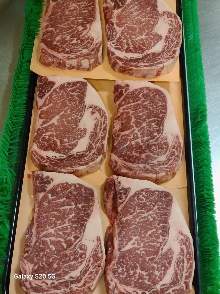 Six marbled raw steaks arranged in a green tray on tan paper.