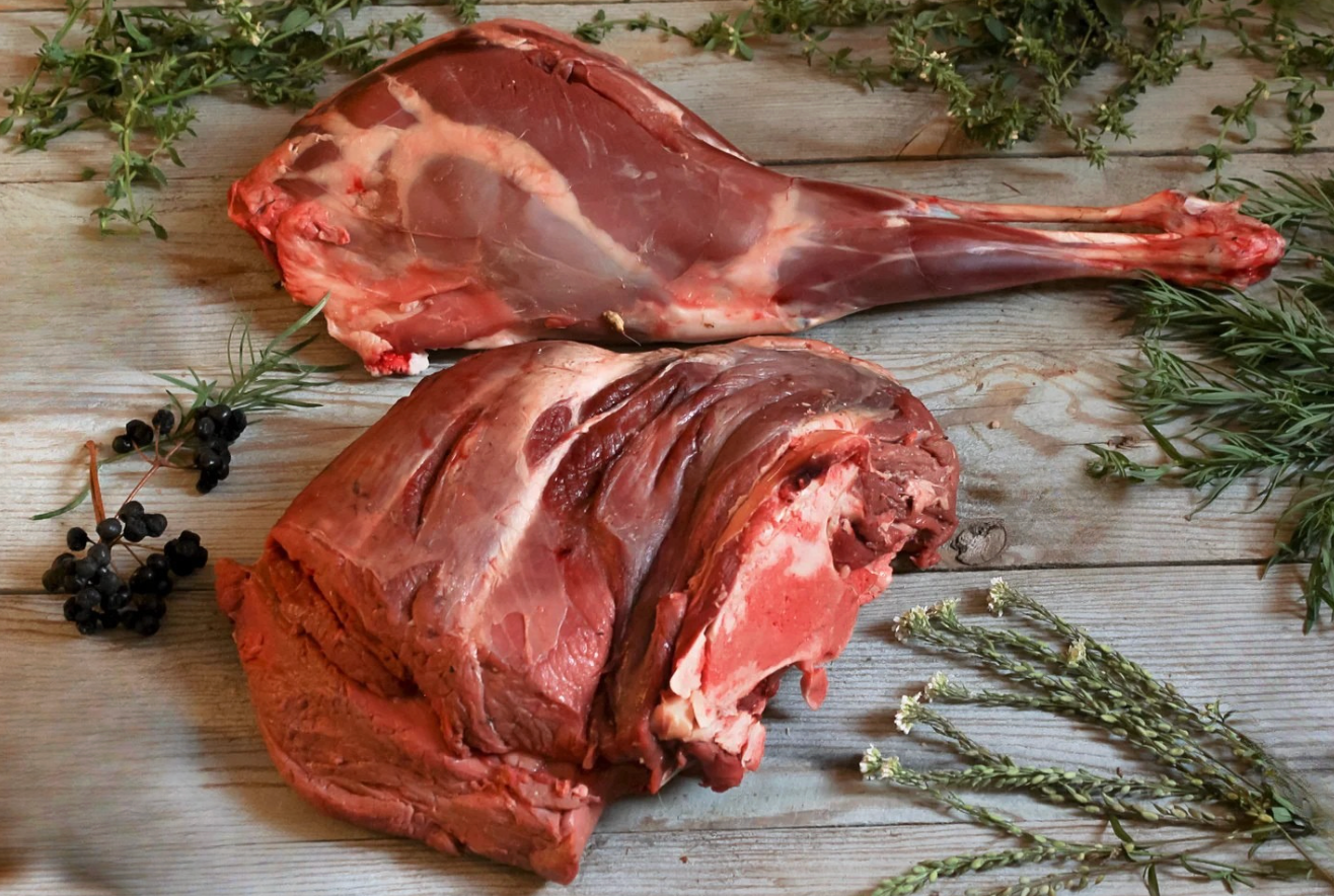 Two raw cuts of red meat on a wooden board with rosemary and berries surrounding them