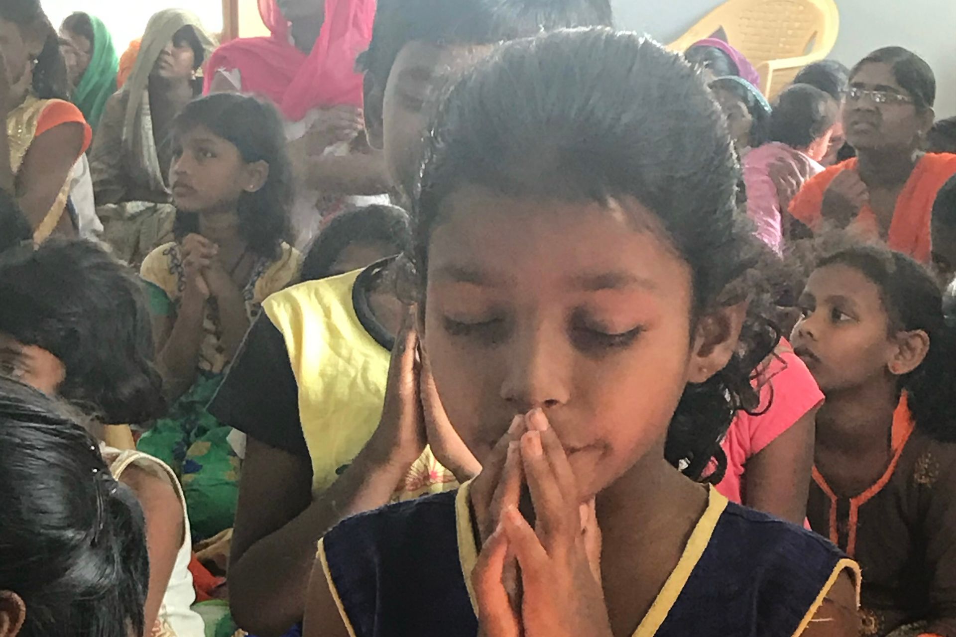 Girl with hands together, eyes closed, praying with others in a room.