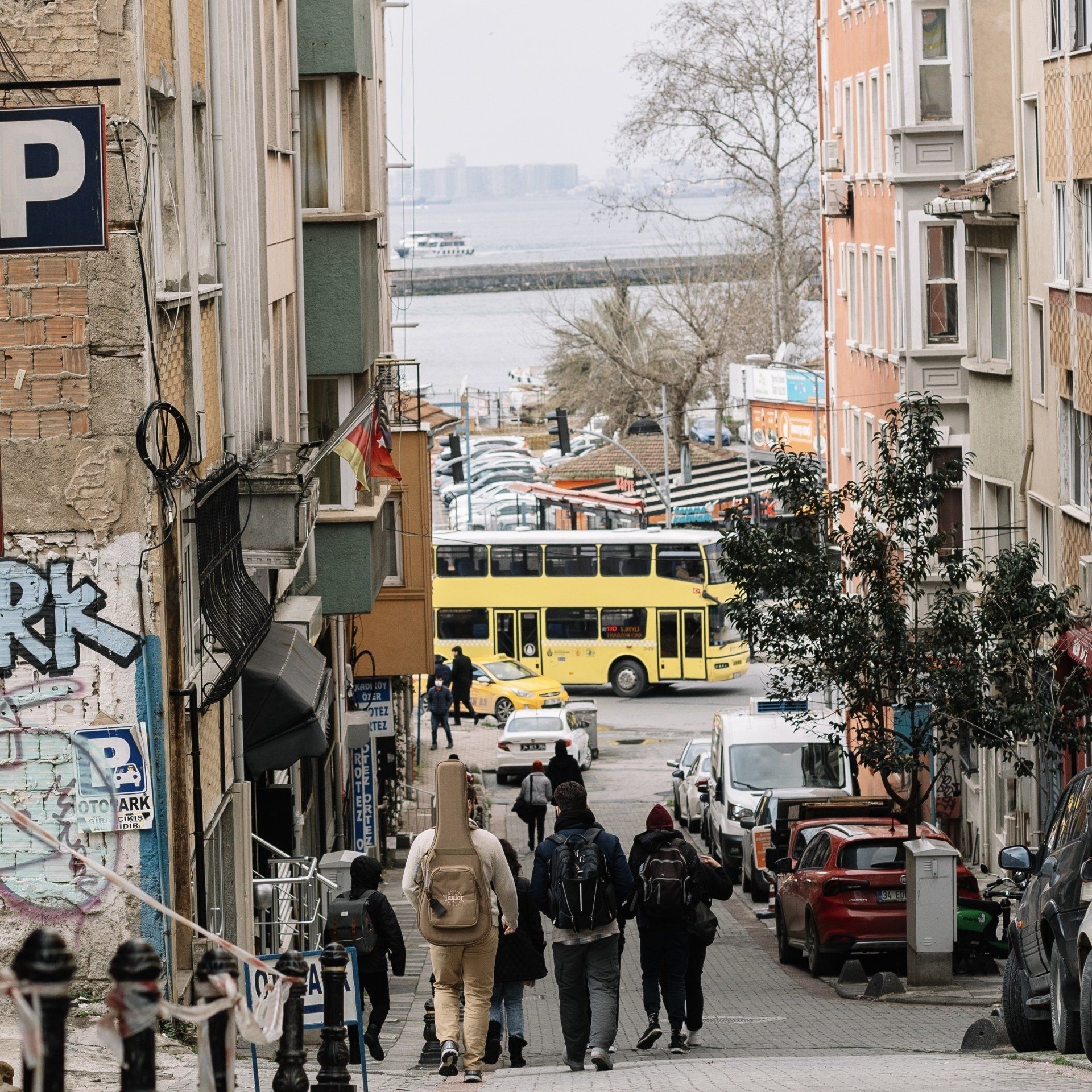 People walk down a sloping city street with a double-decker bus. Buildings line the sides; water visible in the background.