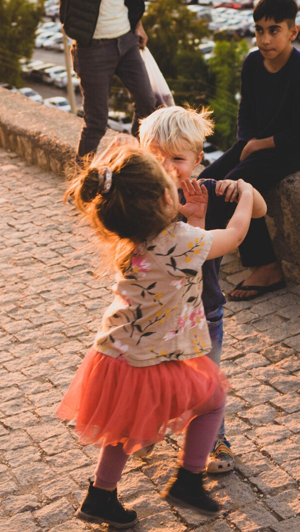 Two children dancing on a stone path; girl in pink tutu and floral top, boy in blue shirt.