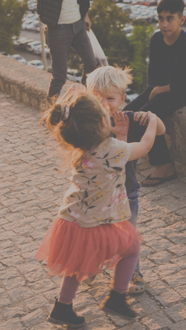 Two children dancing on a stone patio, girl in pink skirt, boy in blue shirt, background with people and cars.