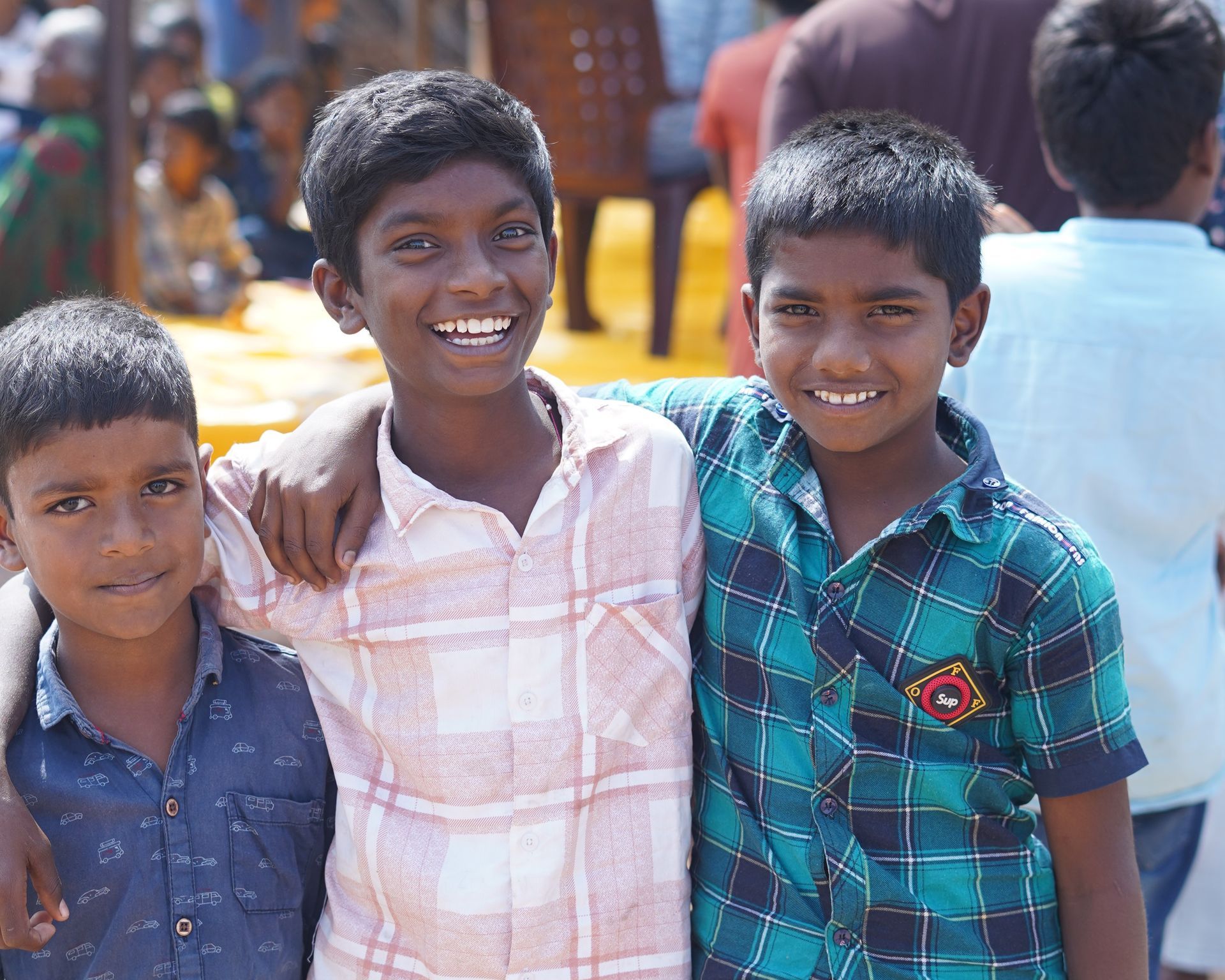 Three smiling boys with arms around each other, wearing button-down shirts, outdoors.