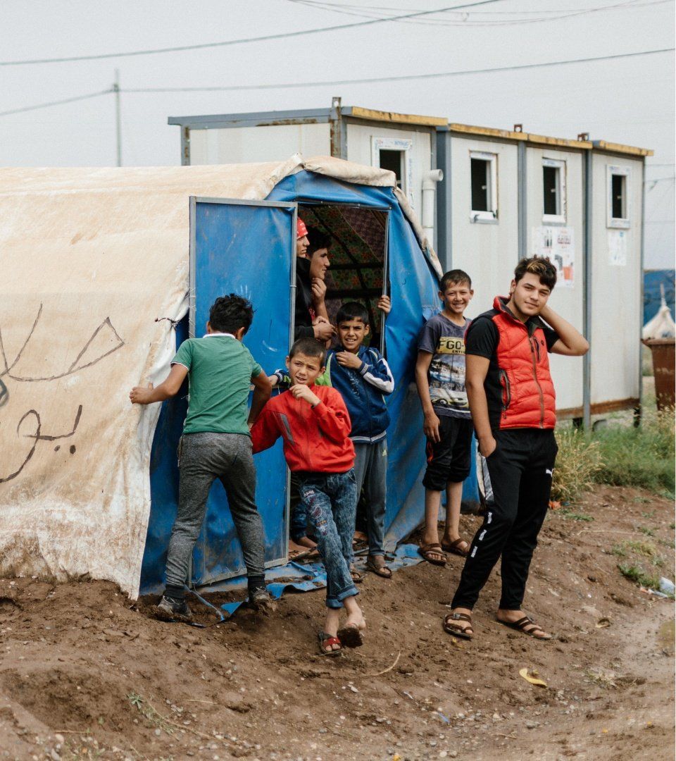 Children outside a blue tent, another structure in the background. Boys are smiling and looking at the camera.