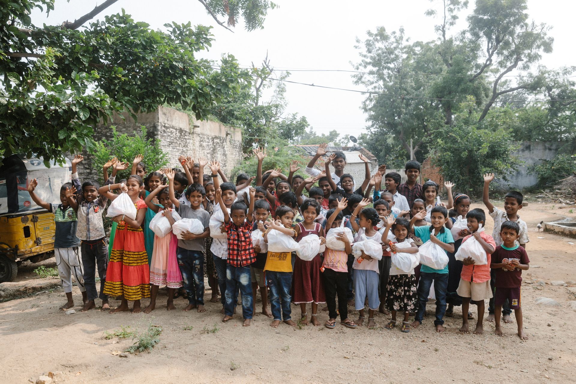 Group of children holding bags, waving, and smiling outside a building in a rural setting.