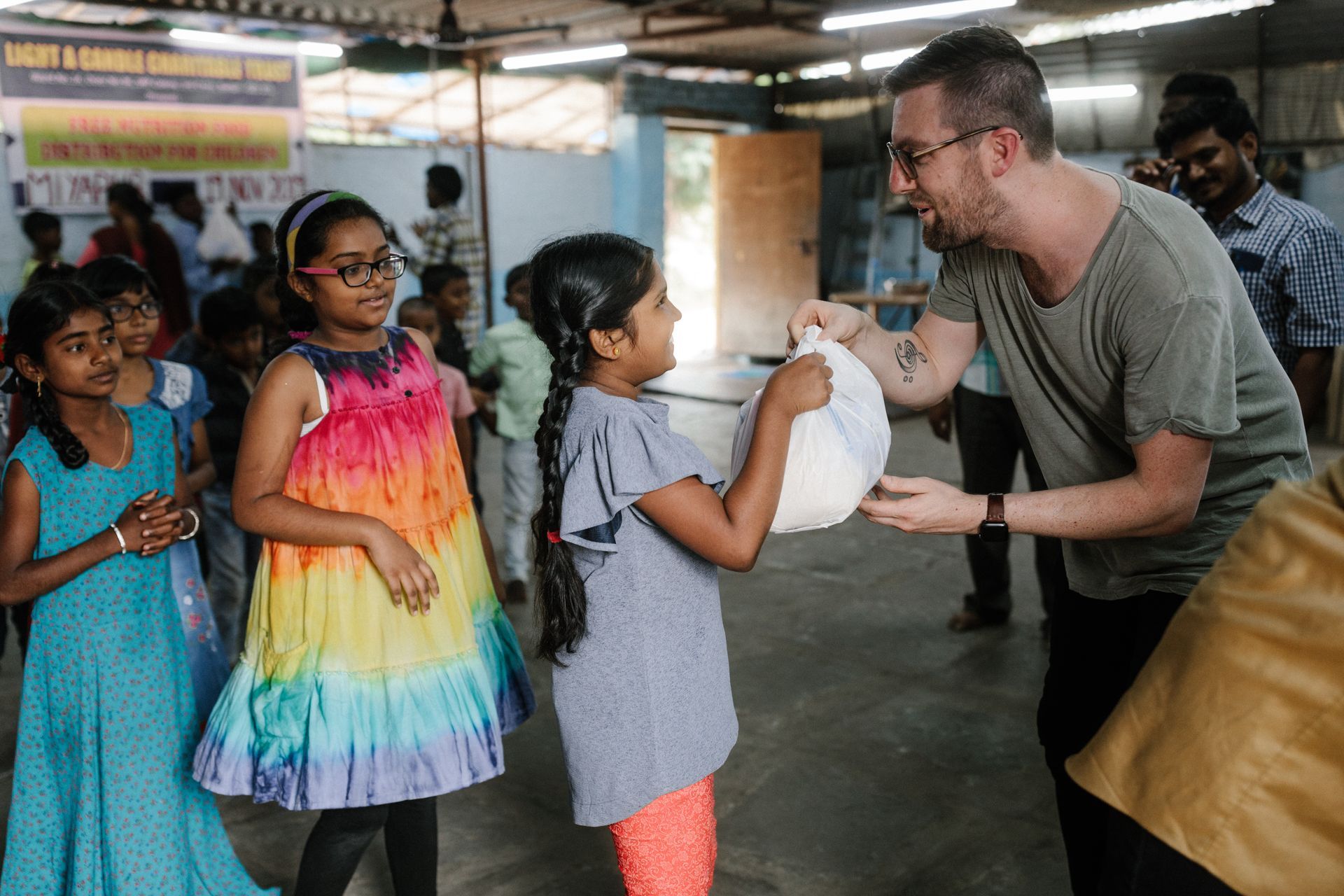Man giving a bag to a girl surrounded by children in a brightly lit room.