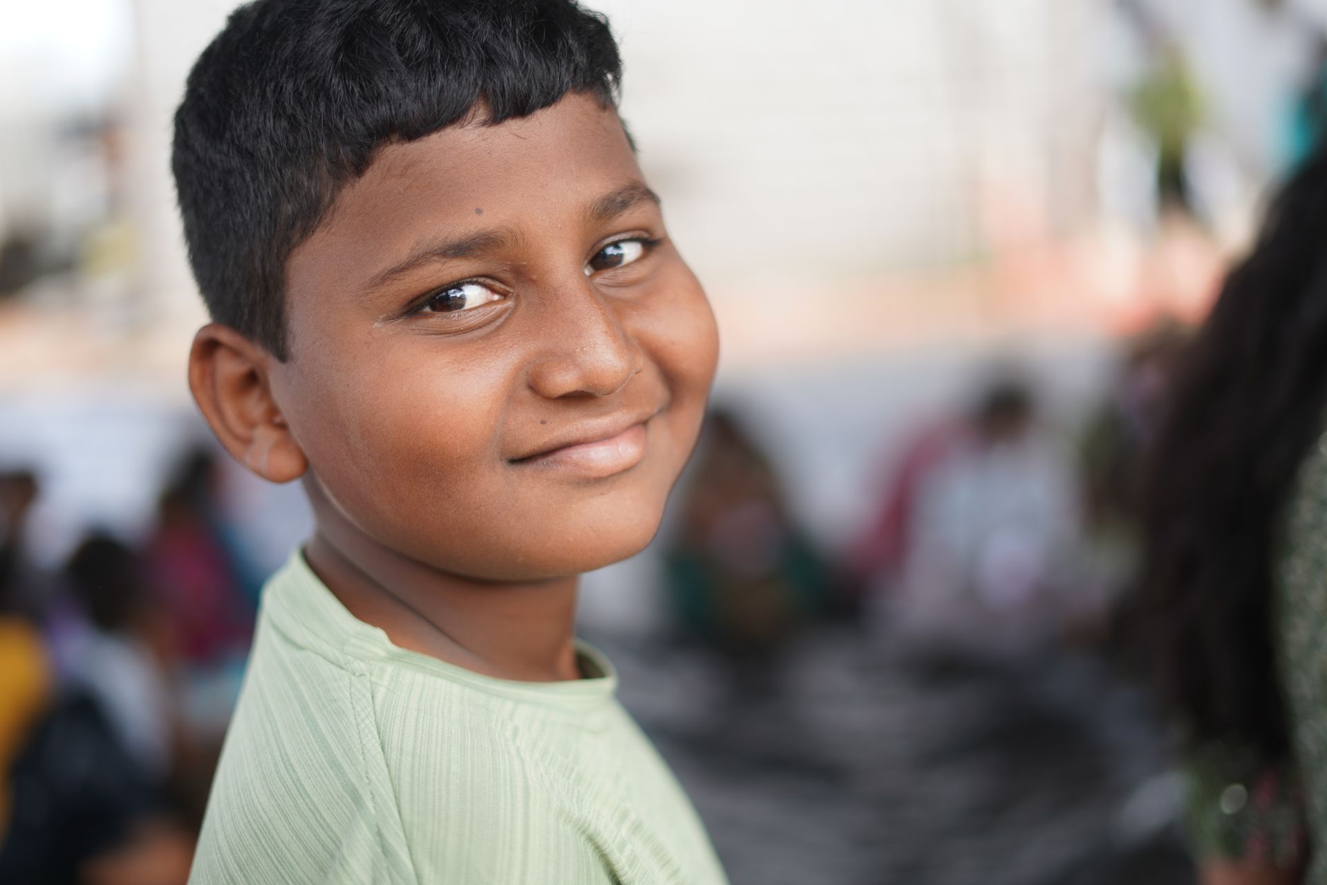 A smiling child looking over their shoulder at the camera, wearing a light green shirt in a blurry outdoor setting.