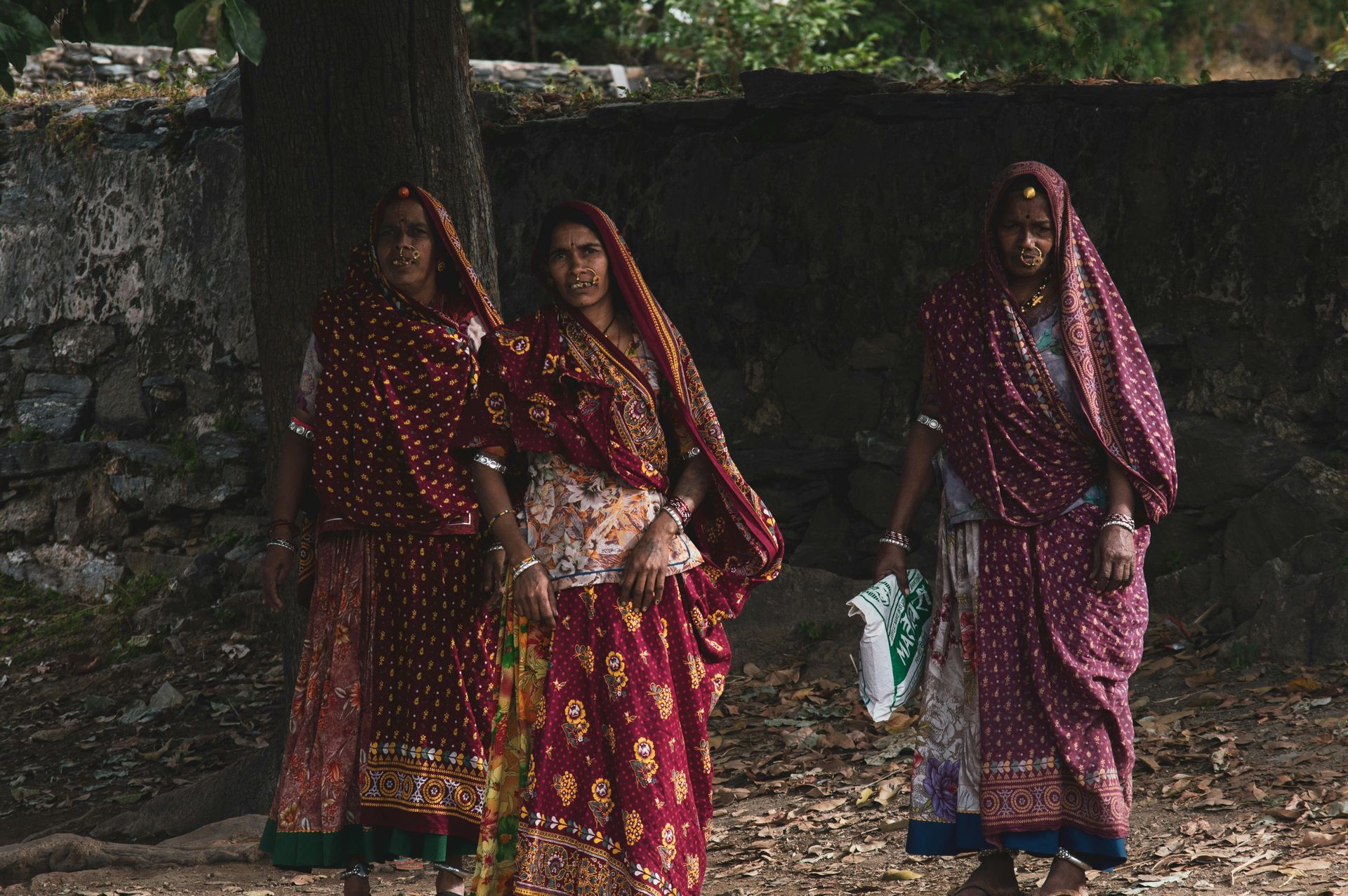 Three women in red patterned saris stand near a tree. One holds a paper.