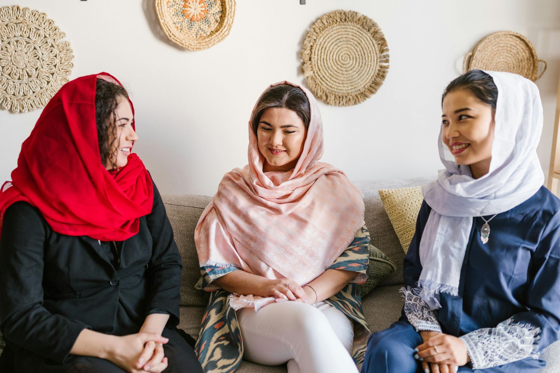 Three women in hijabs sit on a couch, talking. Two wear headscarves, the third a hijab.