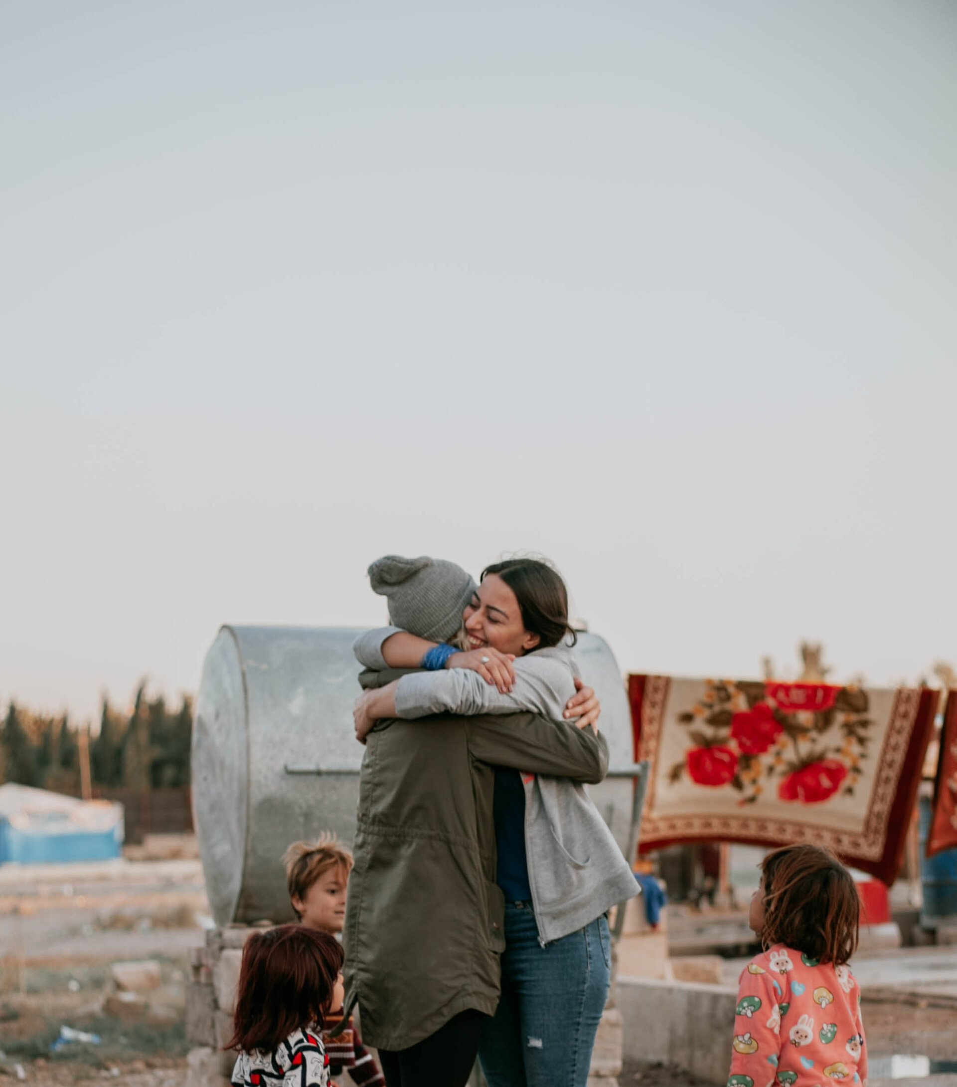 Two people embracing outdoors, surrounded by children and a camp setting, with blankets hanging.