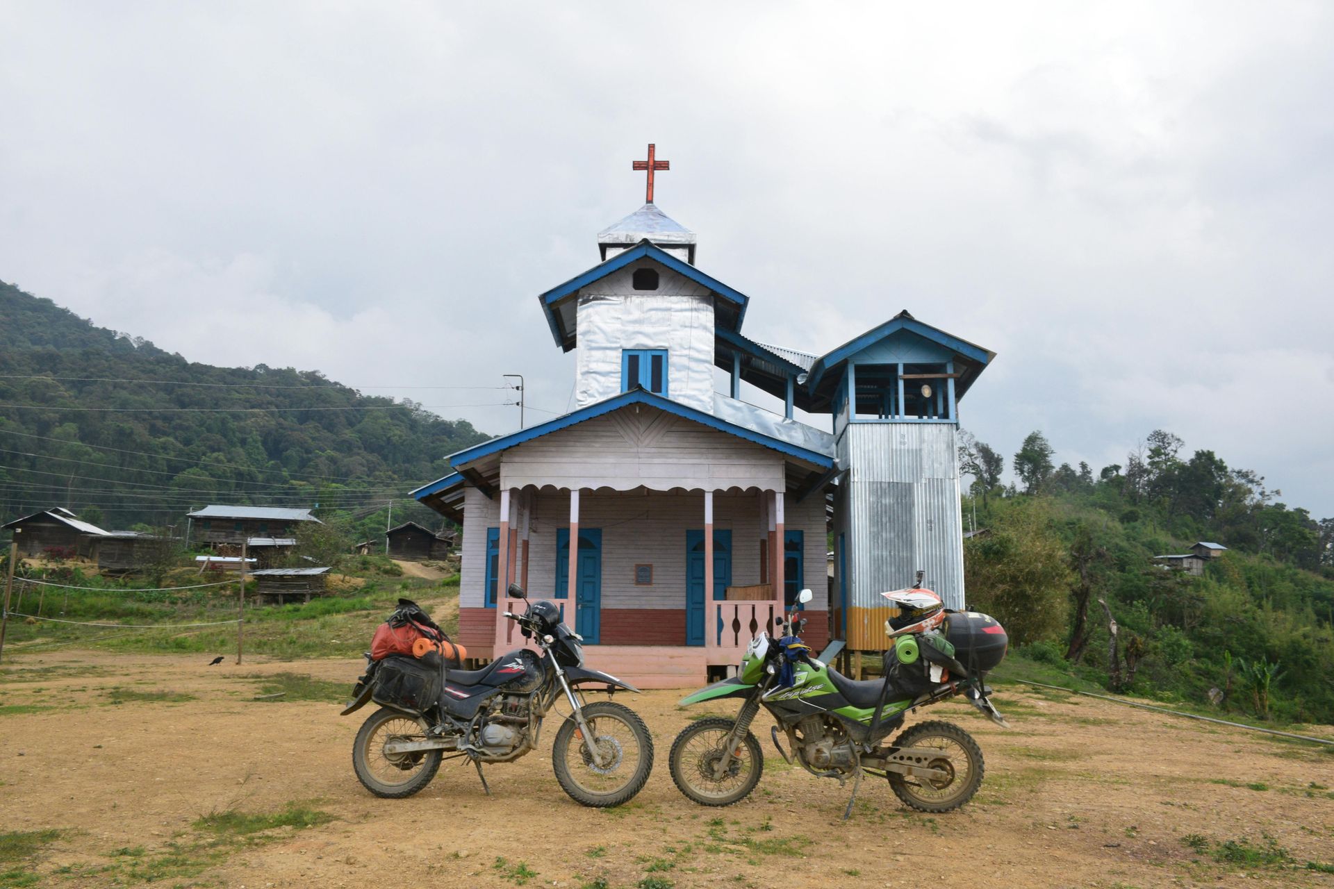 Two motorcycles parked in front of a rural church with a cross on top. Overcast day.
