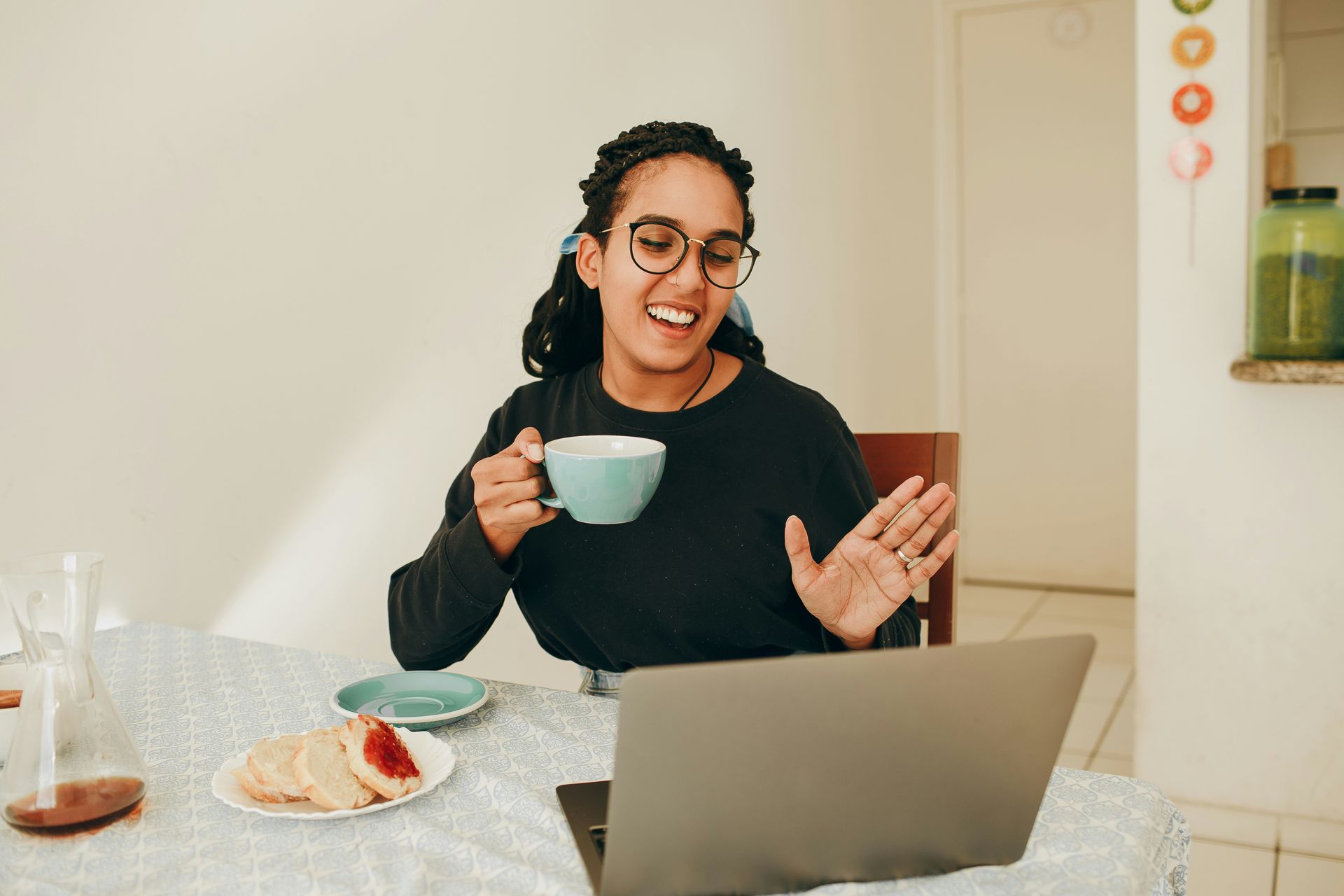 Woman with glasses on a video call, holding a mug. She gestures and smiles, with food and a laptop on the table.