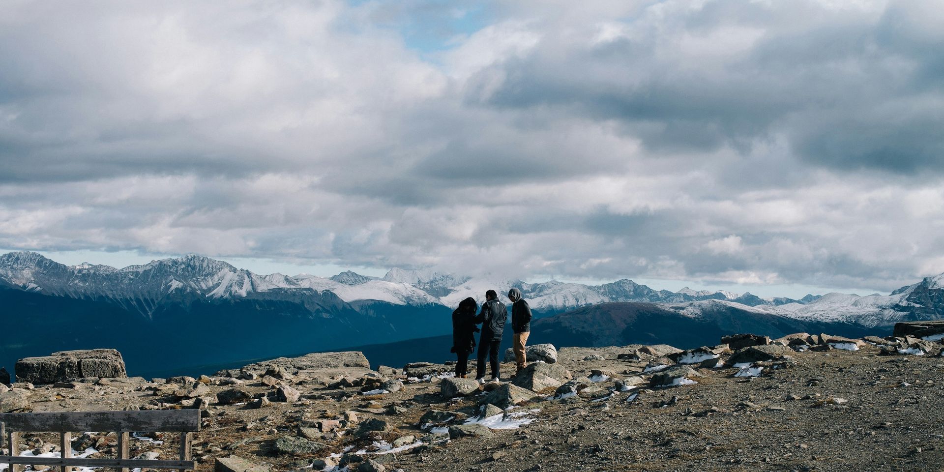 Three figures stand on a rocky summit, gazing at snow-capped mountains under a cloudy sky.
