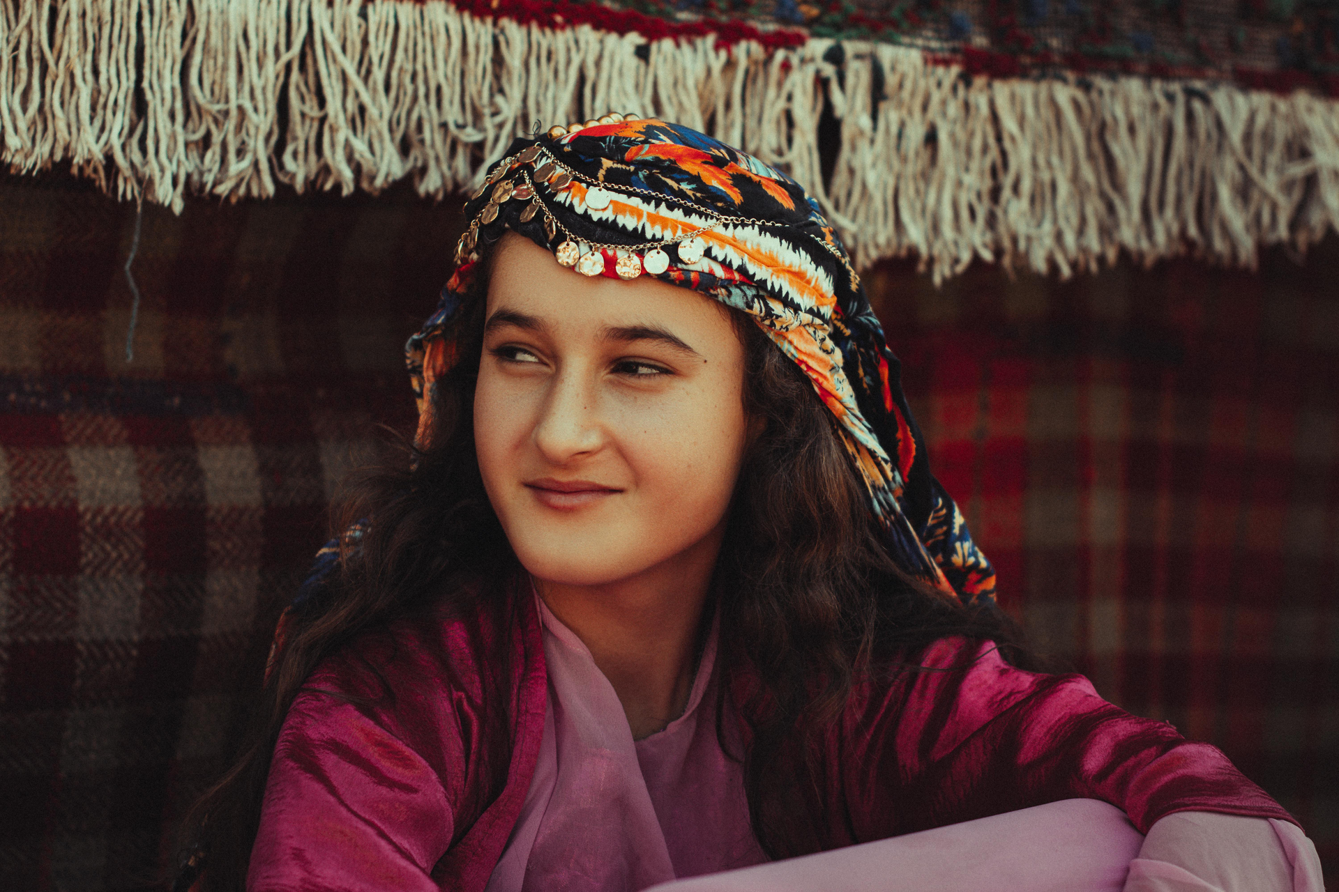Woman wearing a colorful headscarf and pink top, smiling softly. Set against a fringed fabric background.