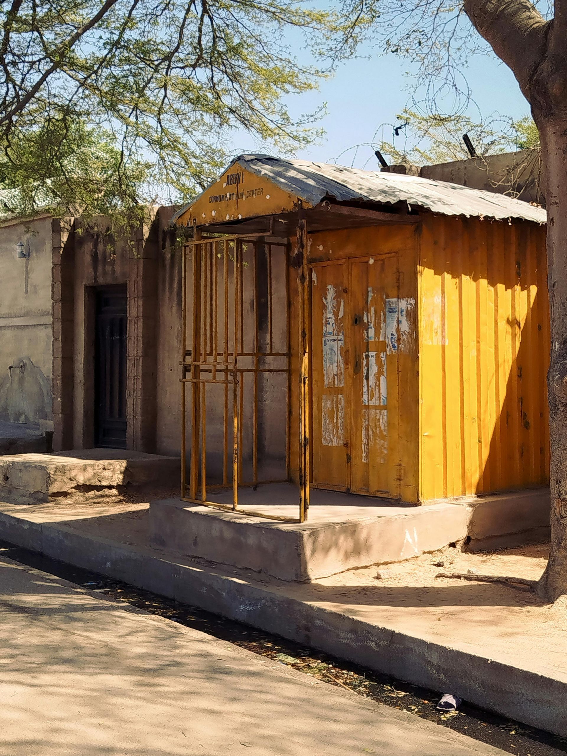 A yellow corrugated metal kiosk with a small porch sits beside a concrete building under a tree.