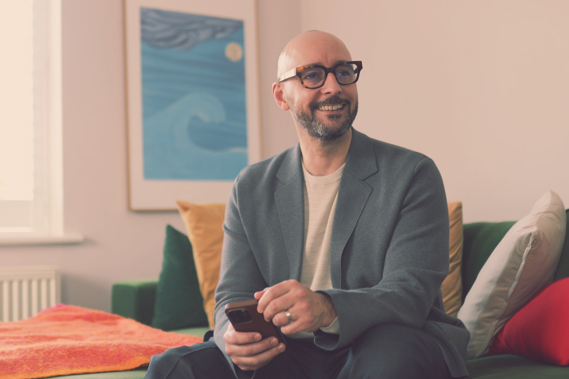 Justin Tate of The Exec Memo with glasses, holding a phone, smiles while seated on a green couch in a living room.