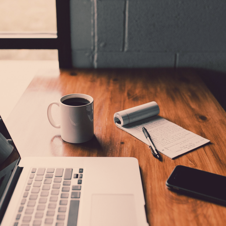 Laptop, mug of coffee, notepad, pen, and phone on wooden table.