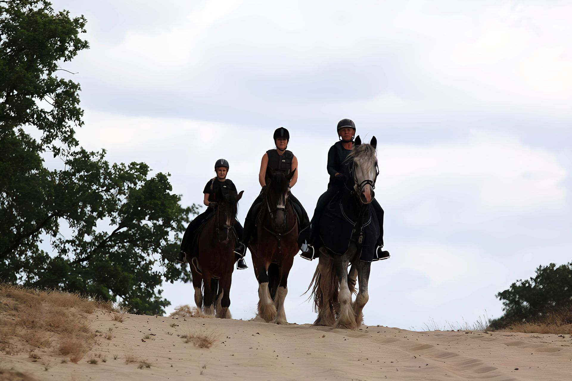 Drie mensen rijden te paard over een zandpad onder een bewolkte hemel.