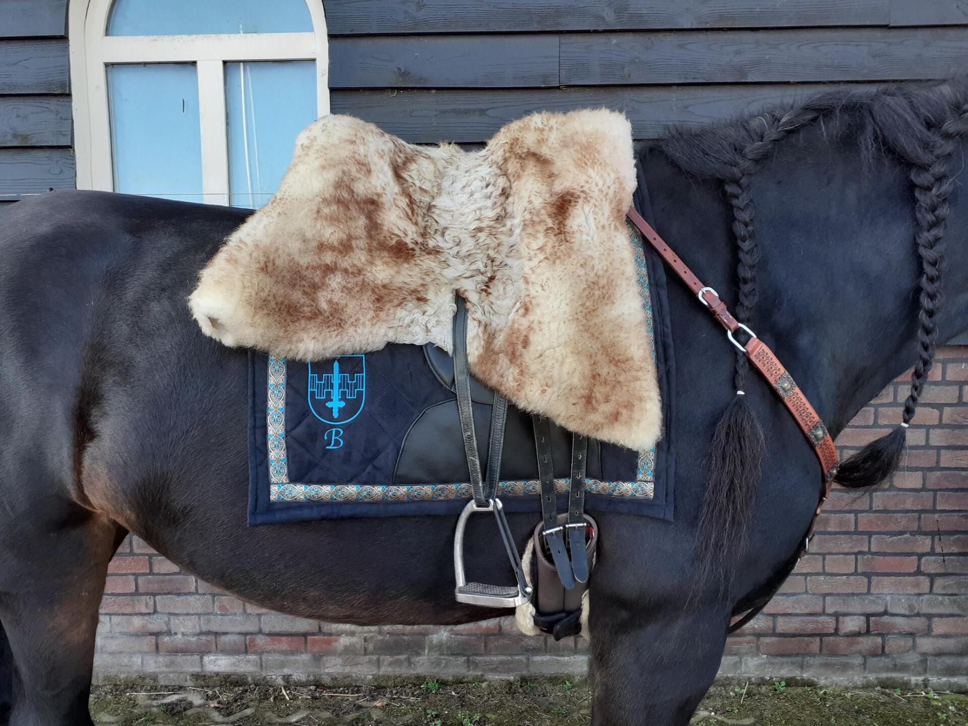 Zwart paard met schapenvacht zadeldek en hoofdstel tegen een houten muur en bakstenen achtergrond.