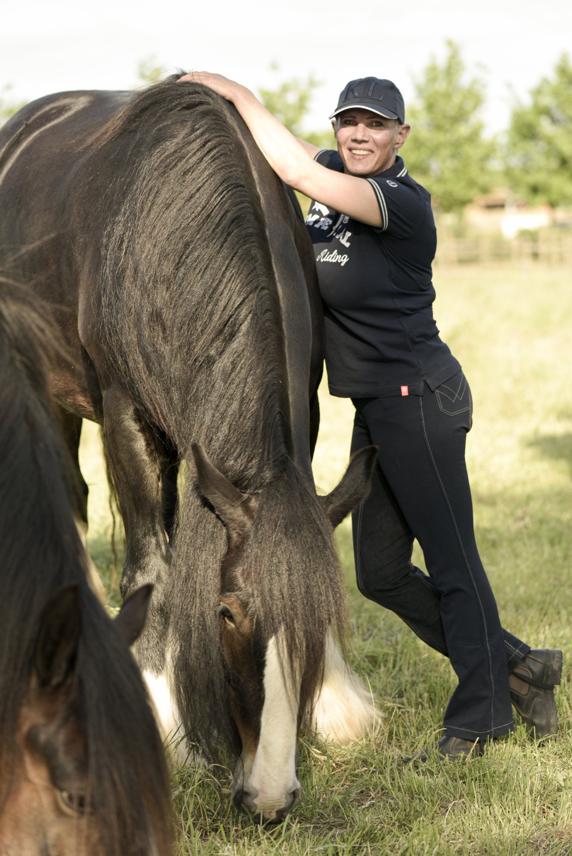 Een vrouw met een pet en donkere kleding leunt tegen een donkergekleurd paard dat graast in een grasveld.