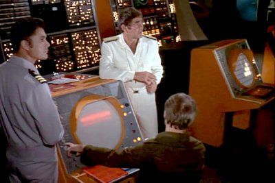 two men in military uniforms are standing next to a man monitoring a console.
