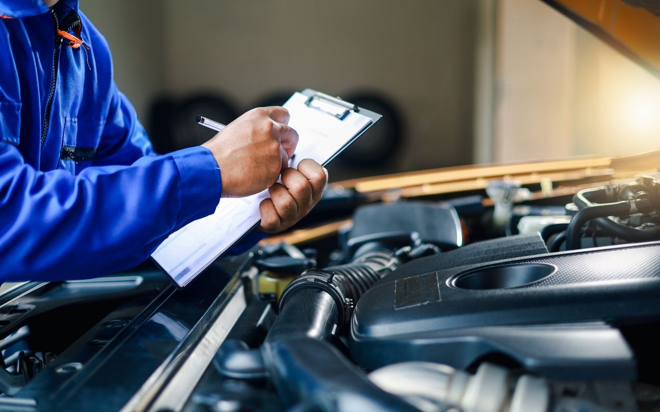 Mechanic in blue uniform inspecting a car engine with a clipboard in a garage
