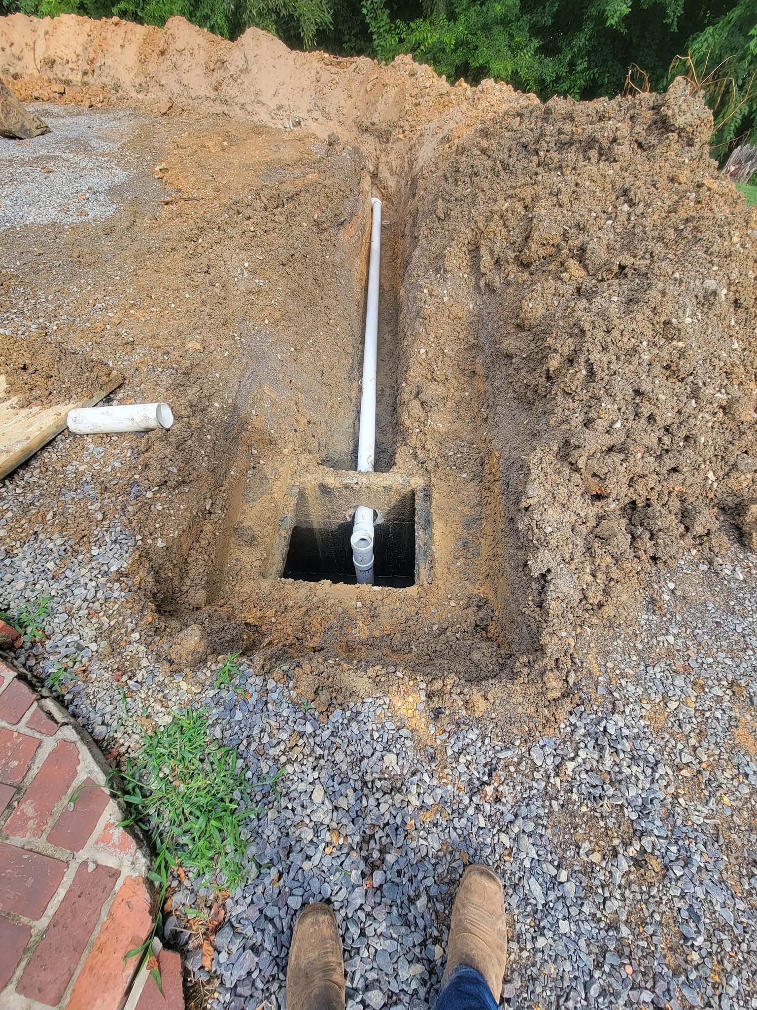 Trench with a white pipe running to a square hole in gravel beside a brick walkway and boots.