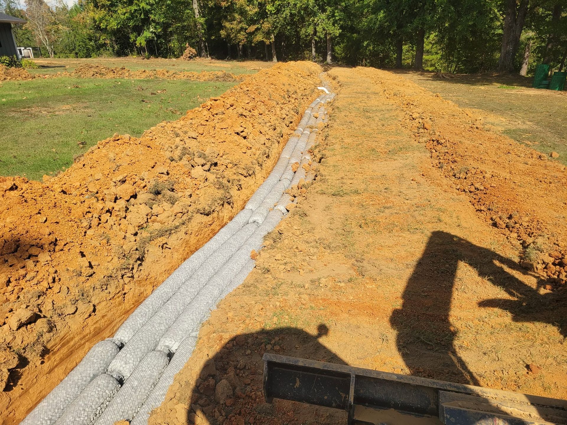 Freshly piled orange soil beside a gray drainage trench at a construction site, with an excavator shadow.